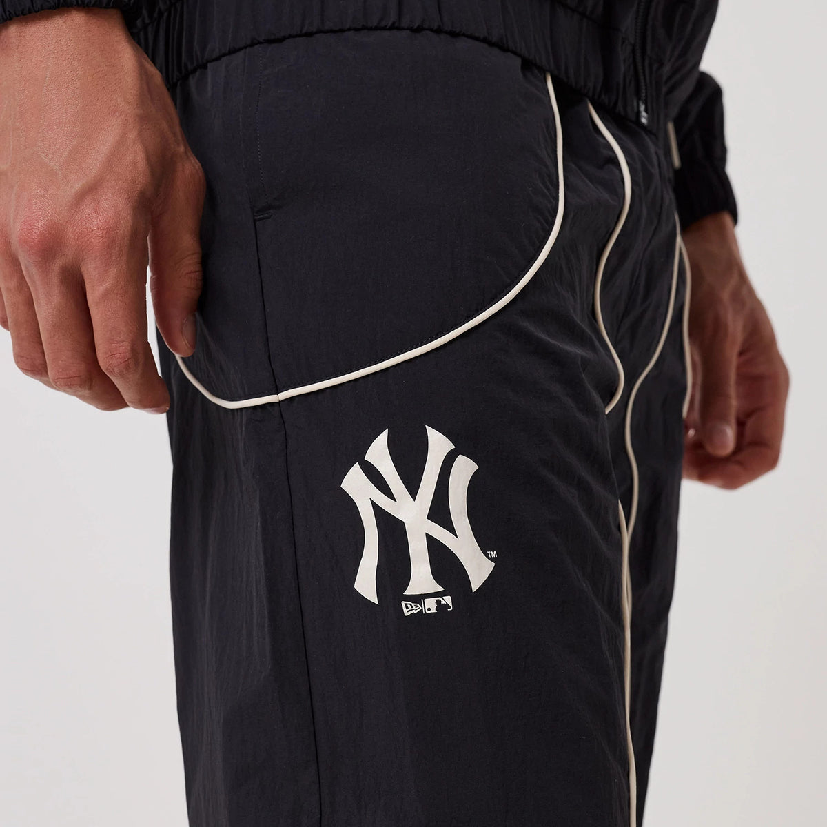 New Era New York Yankees MLB Piping Navy Track Pants, side view showing the white Yankees logo printed on the upper thigh, white piping along the side seam, and gathered ankle cuffs displaying a and sporty style.
