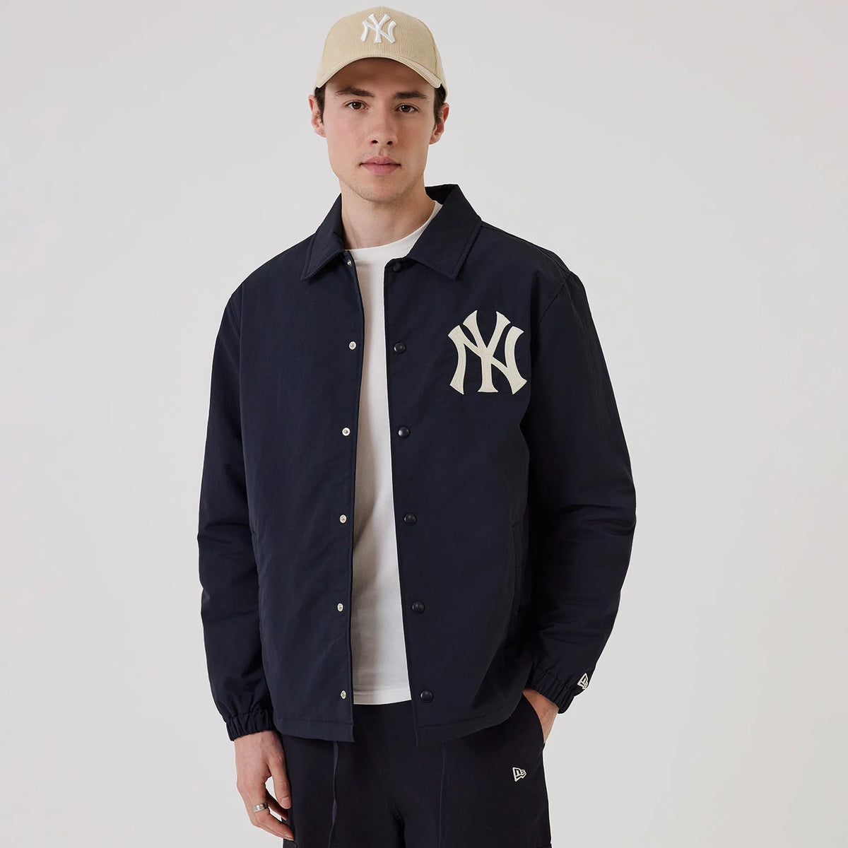 New Era New York Yankees MLB Navy Coach Jacket, front view on-model showing the navy jacket with a white embroidered Yankees logo on the left chest, button-up fastening, and elastic cuffs, styled with a light beige Yankees cap and dark trousers.