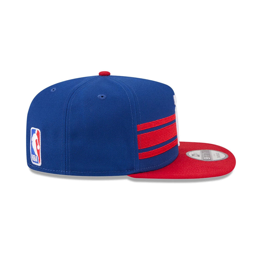 NBA Logo NBA Tip Off 2026 Blue New Era 9FIFTY A-Frame Snapback Adjustable Cap, side view showing the embroidered NBA logo on the back, red contrasting visor, and horizontal red stripes on the structured blue crown.
