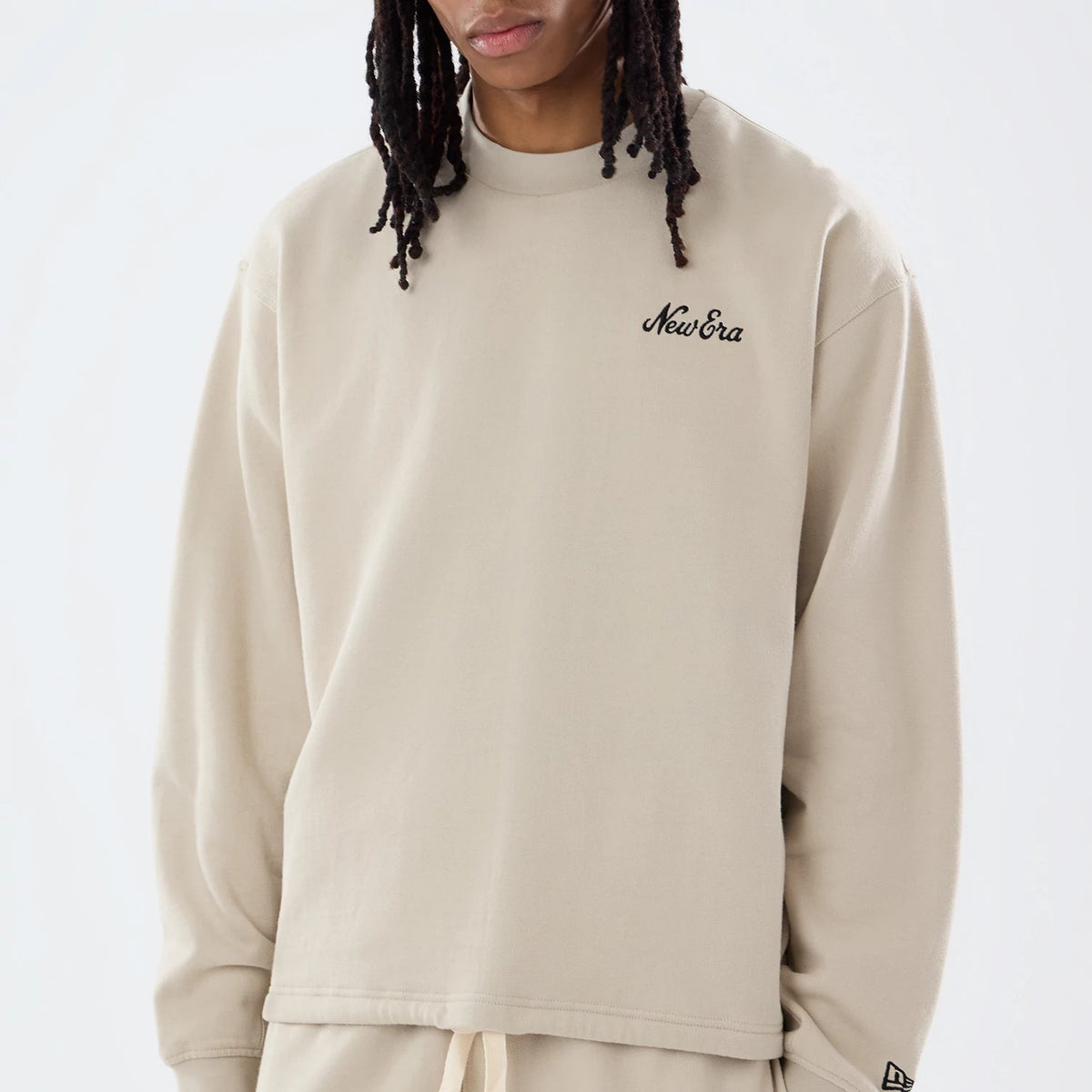 New Era Open Hem Light Beige Oversized Crew Neck Sweater, front view on a model showing the embroidered New Era script logo on the left chest, relaxed drop shoulders, and the open hem design with a New Era flag logo on the left sleeve cuff.