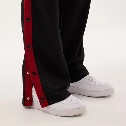 New Era Miami Heat NBA Hardwood Classic Straight Black Straight Leg Joggers, on-model side view showing black joggers with red side stripes and snap button closures, falling over white trainers against a plain background.