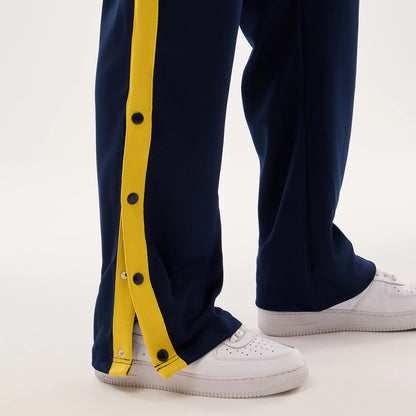 New Era Warriors NBA Hardwood Classic Straight Dark Blue Straight Leg Joggers, close-up view showing dark blue fabric with a contrasting yellow side stripe, black snap buttons along the leg, and white trainers at the base.