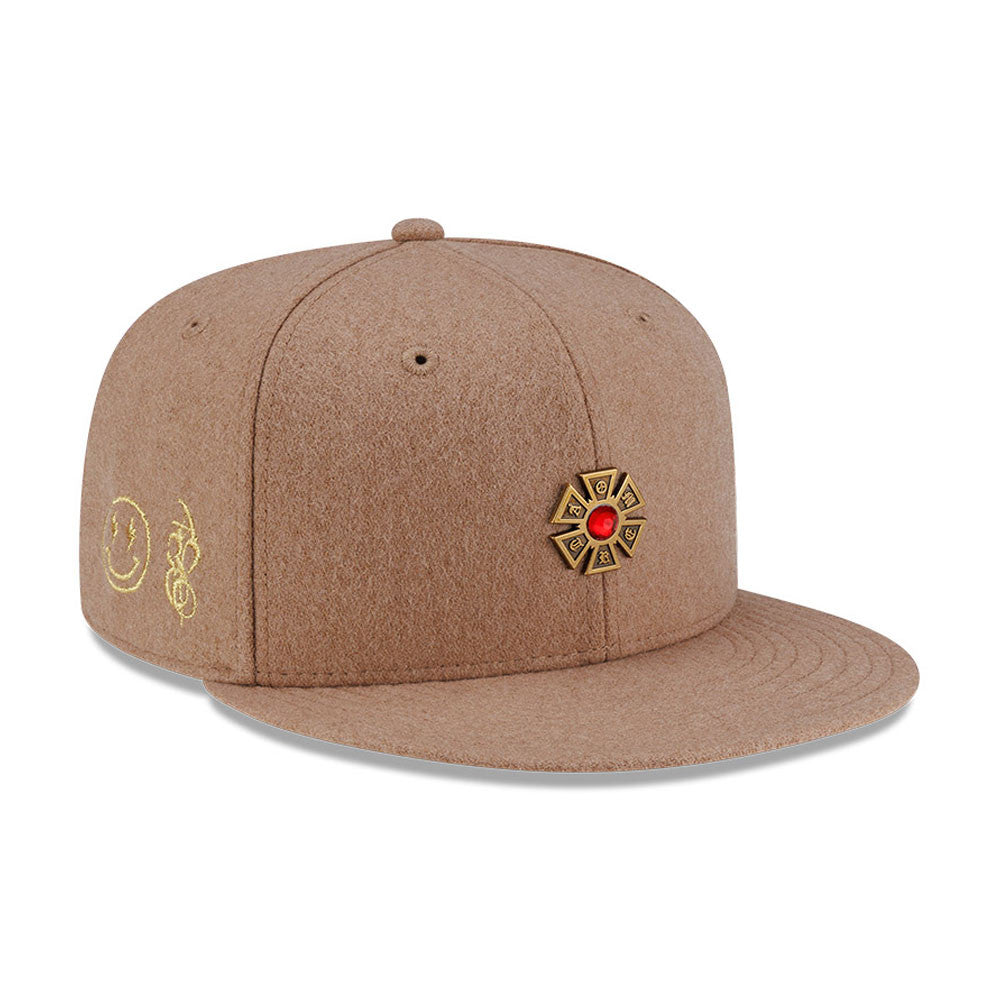 New Era X Omerta Balvin Beige New Era 59FIFTY Fitted Cap, three-quarter view showing a gold cross emblem with a red jewel on the front panel, gold embroidered smiley and logo on the side, and a flat beige visor.
