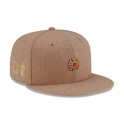 New Era X Omerta Balvin Beige New Era 59FIFTY Fitted Cap, three-quarter view showing a gold cross emblem with a red jewel on the front panel, gold embroidered smiley and logo on the side, and a flat beige visor.