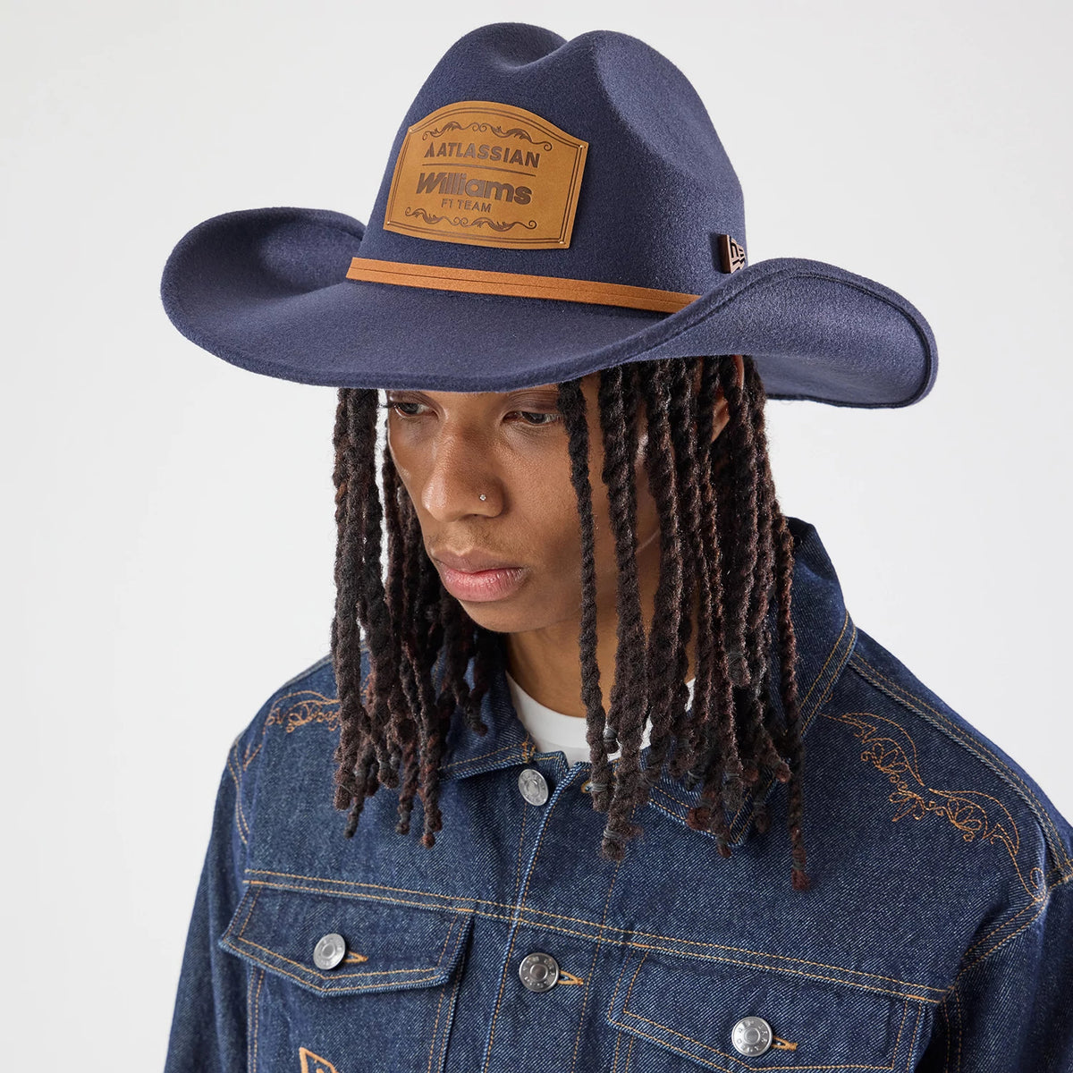 New Era Williams Austin Race Special Navy Wide Brim Fedora Cowboy Hat, on-model view featuring a wide navy visor with a tan leather band above an embroidered Atlassian Williams F1 Team badge, styled with a denim jacket showcasing matching logos.