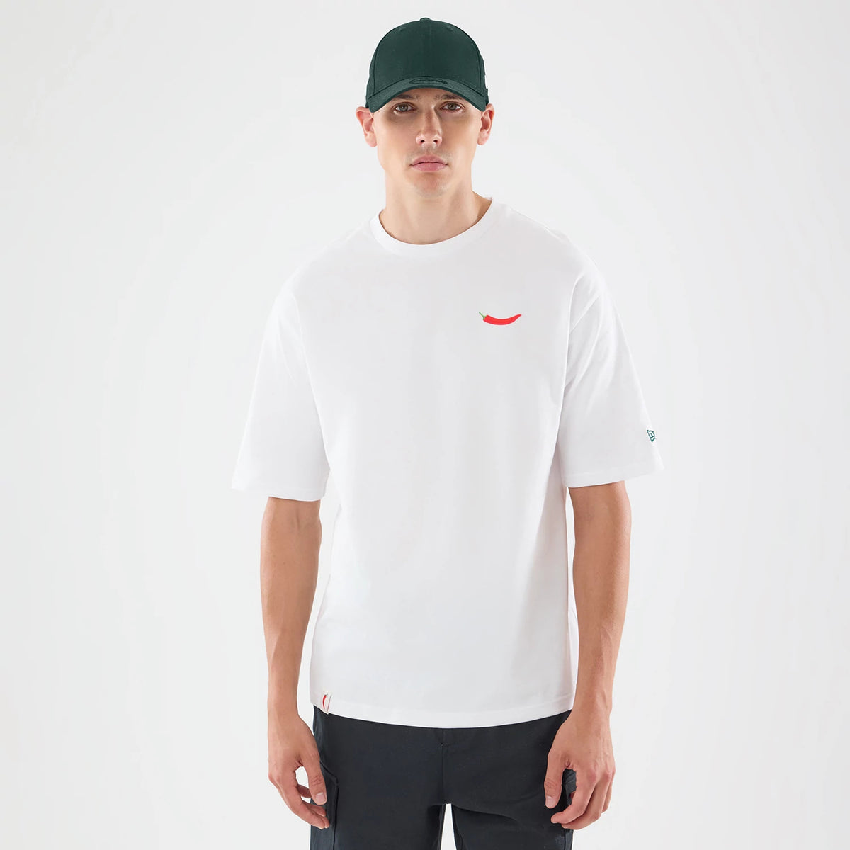 New Era Carlos Sainz Team Fifty5 White Oversized T-Shirt, front view on-model showing a small red printed chilli pepper graphic on the left chest, a relaxed fit with short sleeves, and an embroidered New Era logo on the left sleeve near the cuff.