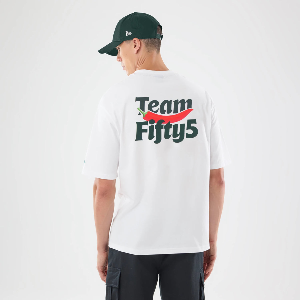 New Era Carlos Sainz Team Fifty5 White Oversized T-Shirt, back view on a model wearing a dark cap, displaying Team Fifty5 graphics centred on the back in black and red, and short sleeves enhancing the relaxed oversized fit.