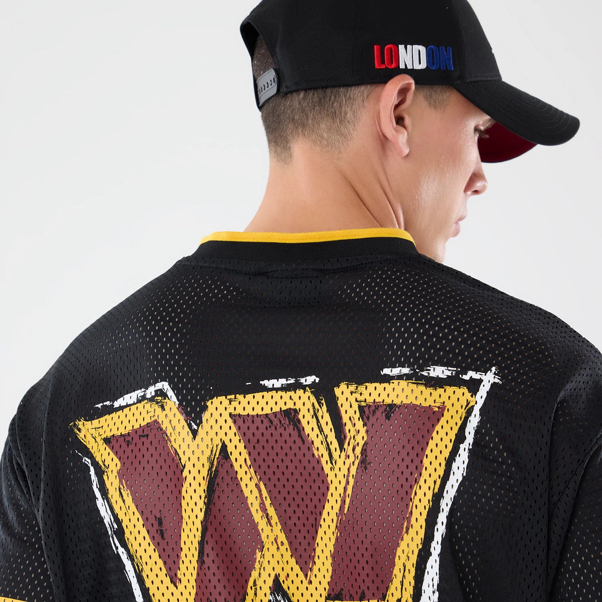 New Era Washington Commanders NFL International Games 2026 Graphic Black Mesh Jersey, back view on a model wearing a black cap, featuring a large distressed yellow and red W logo below the yellow-trimmed neckline on the mesh fabric.