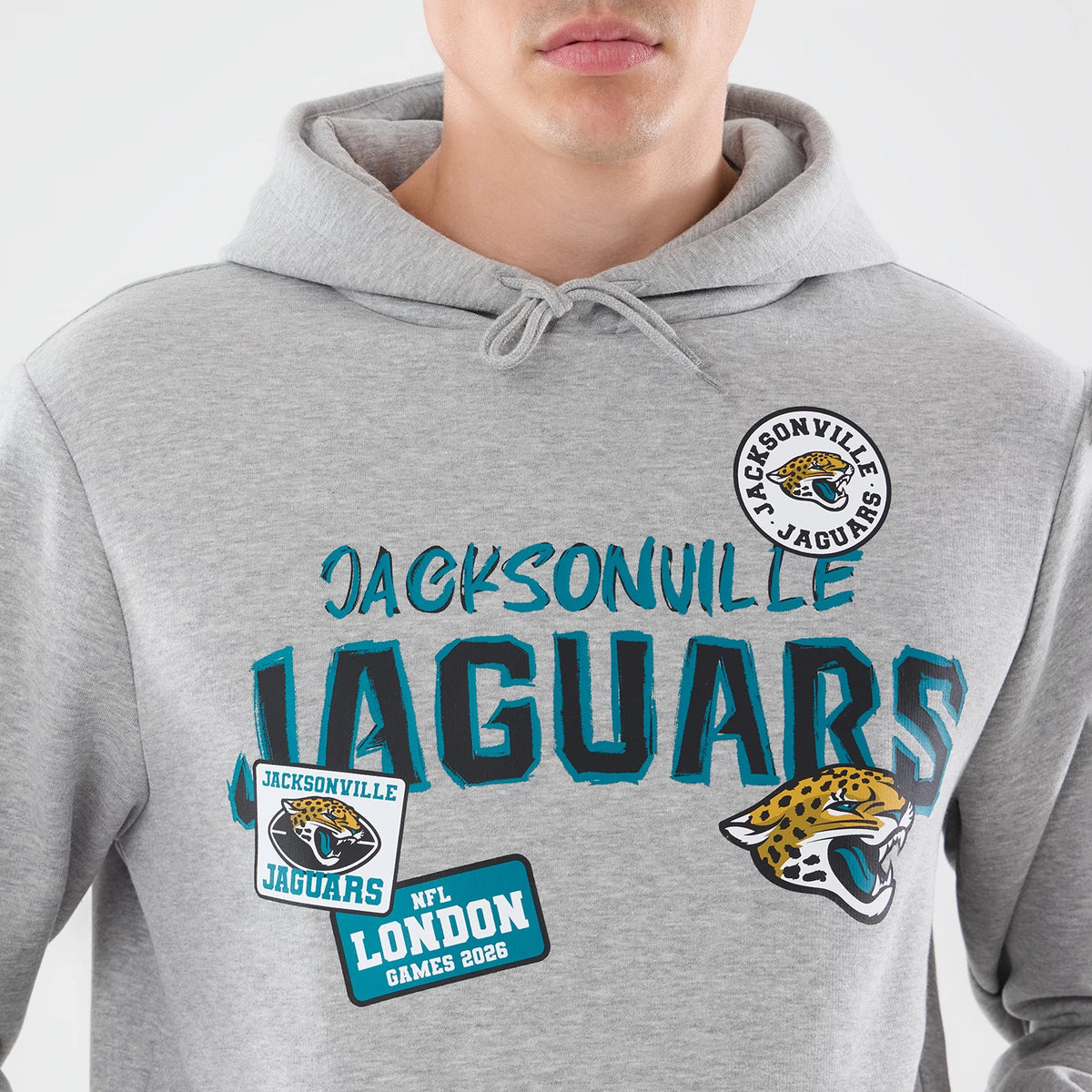 New Era Jacksonville Jaguars NFL Patch Grey Pullover Hoodie, front view on a model showing teal Jacksonville Jaguars printed across the chest, embroidered Jacksonville logo patch above, and additional patches including NFL London Games 2026 below.