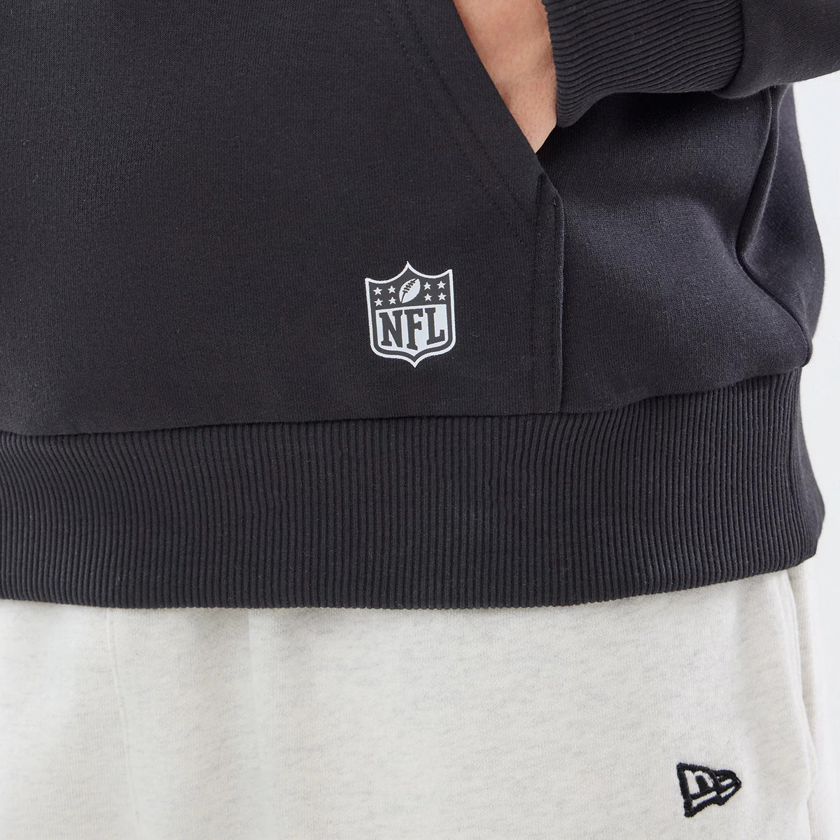 New Era Atlanta Falcons NFL Patch Black Pullover Hoodie, side view highlighting the white embroidered NFL logo near the ribbed hem of the black hoodie, with the New Era logo in black on light grey trousers below.
