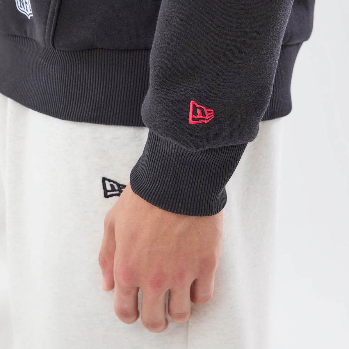 New Era Atlanta Falcons NFL Patch Black Pullover Hoodie, on-model view showing the lower sleeve with a red embroidered New Era logo on the cuff, contrasted against light trousers with a black embroidered New Era logo near the pocket.