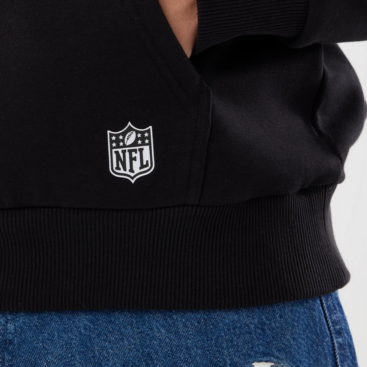 New Era New Orleans Saints NFL Patch Black Pullover Hoodie, front view showing the lower section with a ribbed waistband, a white printed NFL shield logo near the pocket, and styled with blue denim jeans peeking from below.