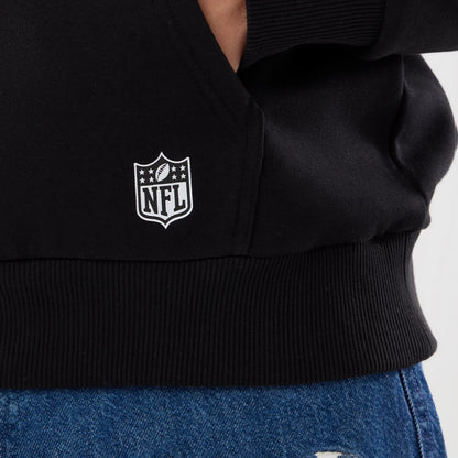 New Era New Orleans Saints NFL Patch Black Pullover Hoodie, front view showing the lower section with a ribbed waistband, a white printed NFL shield logo near the pocket, and styled with blue denim jeans peeking from below.