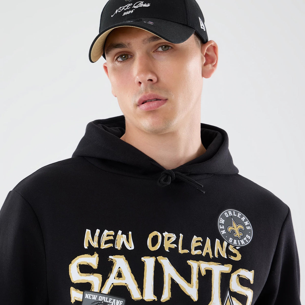 New Era New Orleans Saints NFL Patch Black Pullover Hoodie, front view on a model showing the New Orleans Saints lettering in gold and white on the chest, a round patch with the team logo on the upper chest, and a drawstring hood.