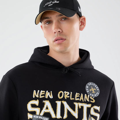 New Era New Orleans Saints NFL Patch Black Pullover Hoodie, front view on a model showing the New Orleans Saints lettering in gold and white on the chest, a round patch with the team logo on the upper chest, and a drawstring hood.