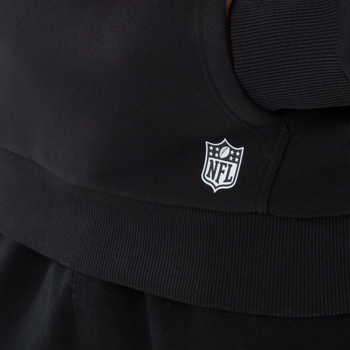New Era Detroit Lions NFL Patch Black Pullover Hoodie, side view showing a small white embroidered NFL shield logo near the ribbed hem, the hoodie’s black fabric, and stitching details around the pocket area.