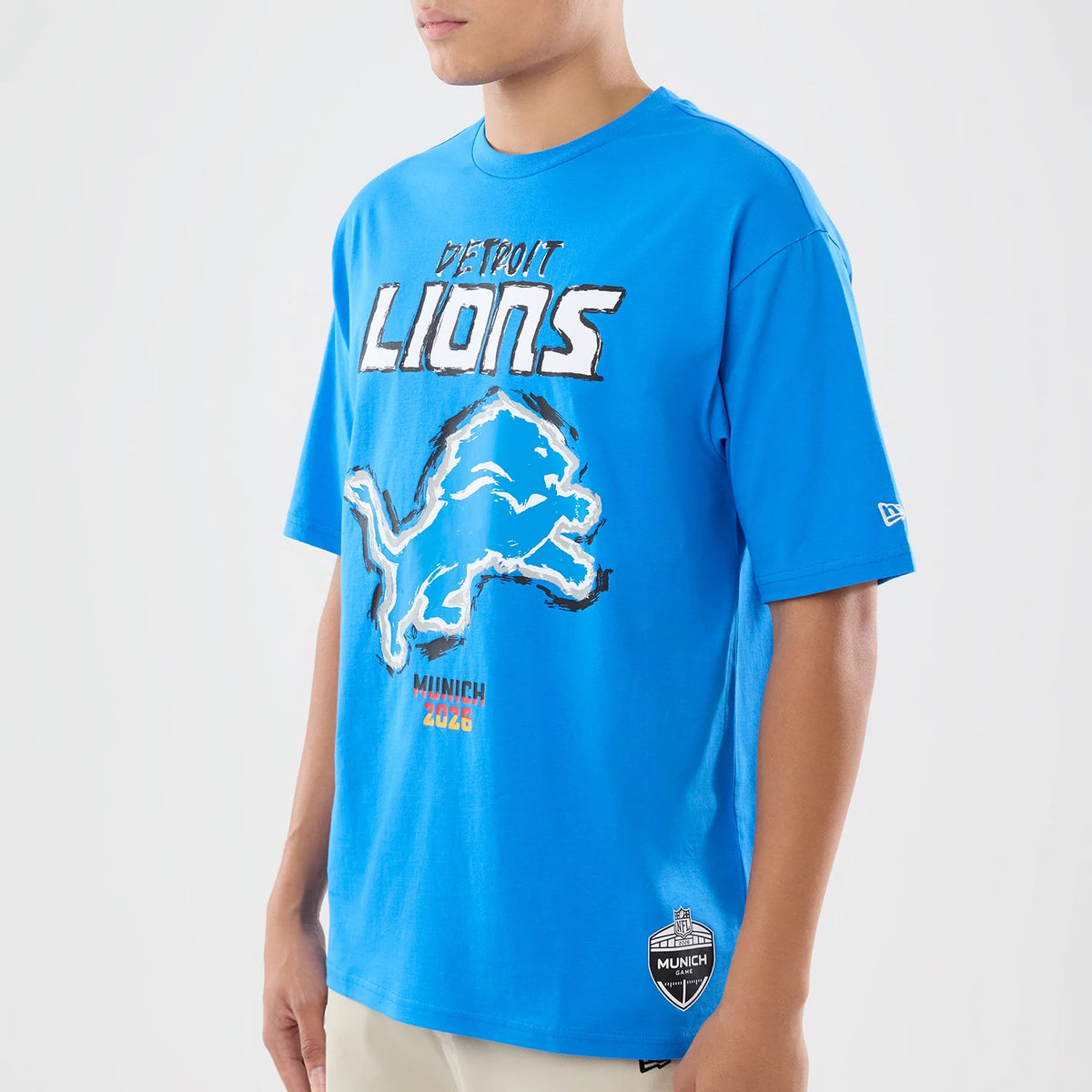 New Era Detroit Lions NFL Graphic Slogan Blue Oversized T-Shirt, front three-quarter view showing the Detroit Lions lettering and large lion graphic on the chest, a small embroidered New Era logo on the sleeve, and a Munich 2023 badge near the hem.