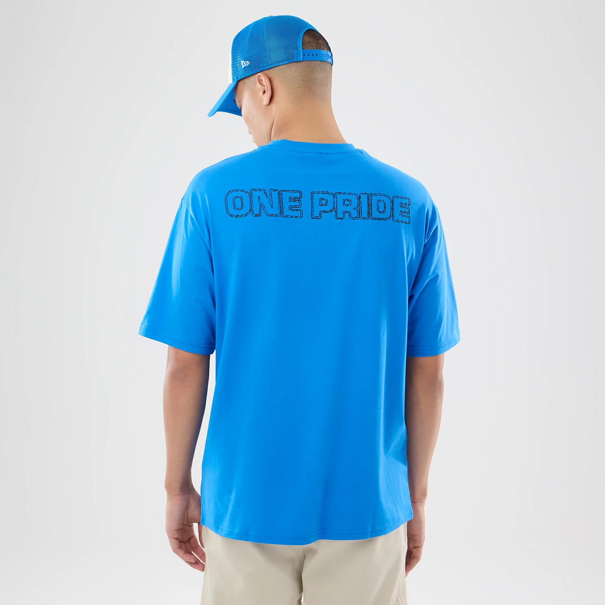New Era Detroit Lions NFL Graphic Slogan Blue Oversized T-Shirt, back view on-model showing prominent One Pride lettering across the shoulders, blue fabric, and paired with a blue cap featuring an embroidered New Era logo on the side.