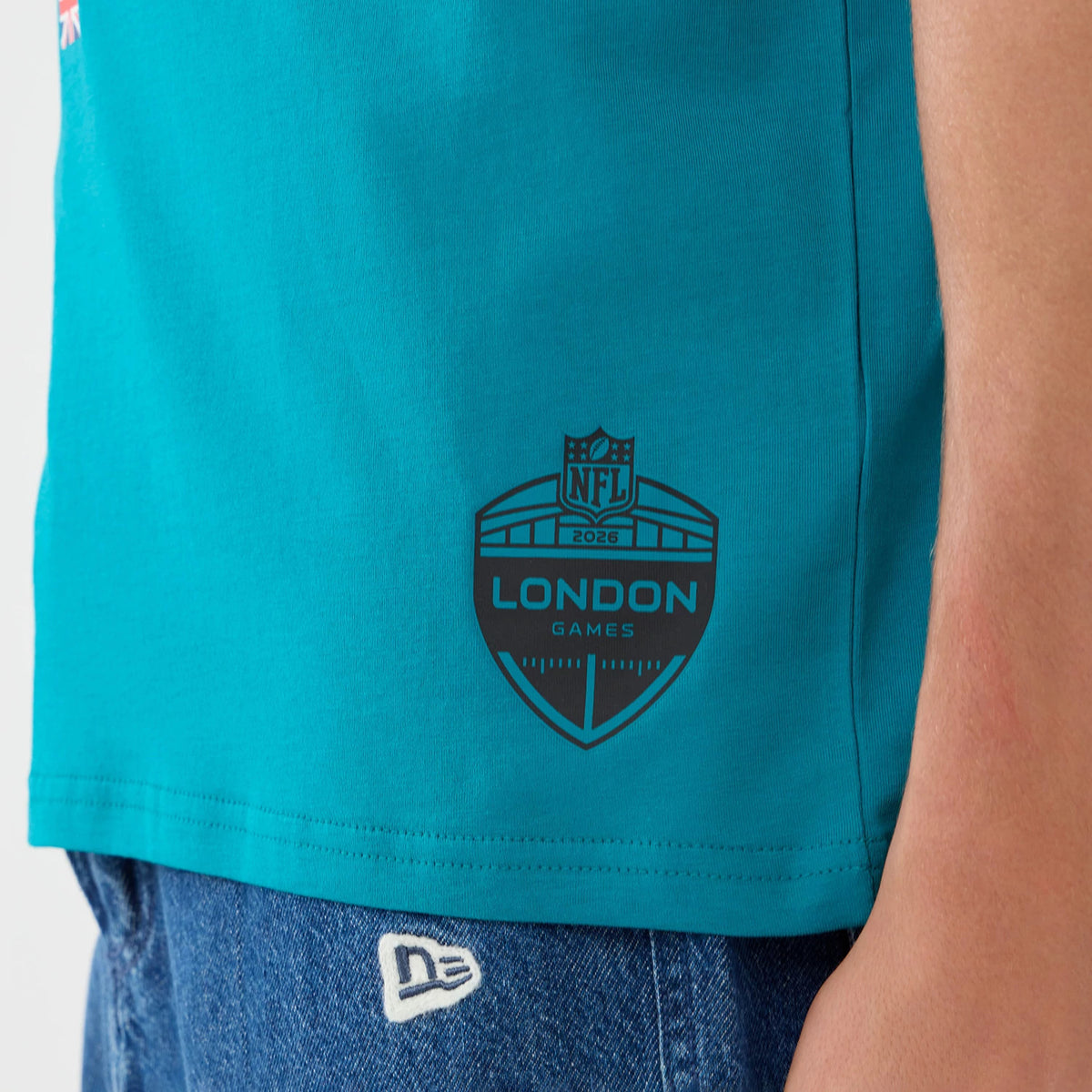 New Era Jacksonville Jaguars NFL Graphic Slogan Turquoise Oversized T-Shirt, side view showing an embossed London Games logo in black near the hem, the turquoise fabric contrasting with blue denim, and the white embroidered New Era logo on the jeans.