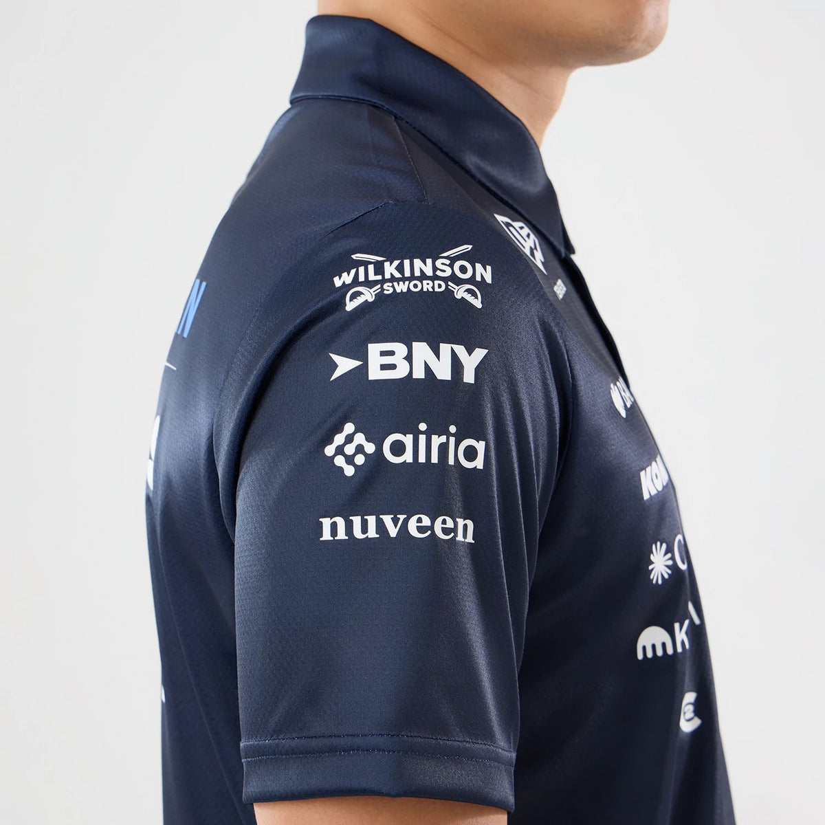 New Era Williams Singapore Navy Tech Polo T-Shirt, side view showing multiple white printed sponsor logos on the sleeve and shoulder, including Wilkinson Sword, BNY, Airia, and Nuveen, with a folded collar.