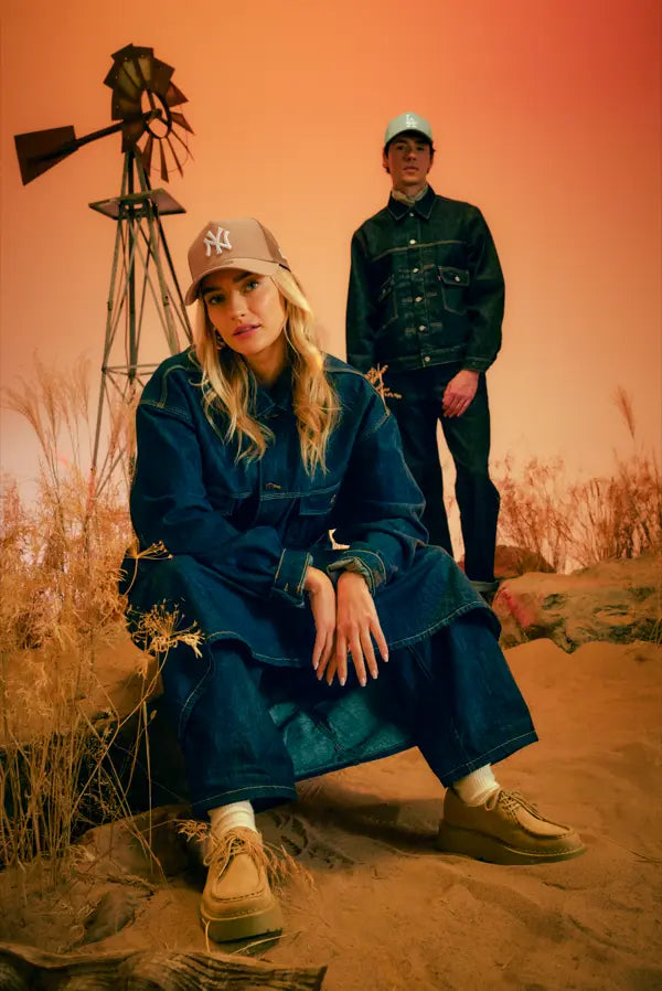 A woman wearing a dark blue denim jacket and matching wide-leg jeans sits on a sandy ridge in a desert landscape, while a man in a dark green denim jacket and pants stands behind her. Both are wearing New York Yankees A-Frame Caps.