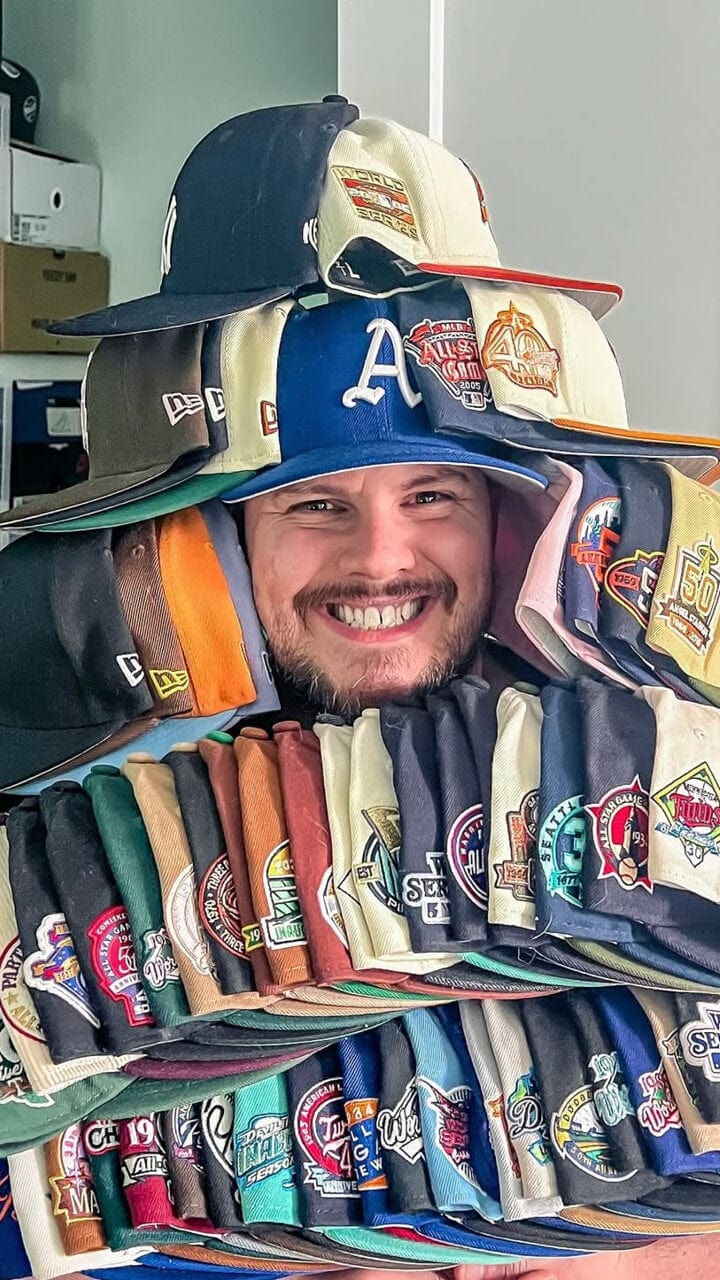 A man smiling with a collection of New Era baseball caps piled around him.