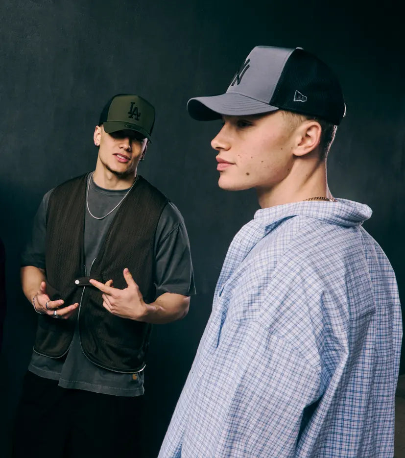 Two young men posing in streetwear against a dark studio background. The model in the foreground wears a blue and white plaid shirt and a grey New York Yankees Trucker E-Frame Cap, while the model behind him wears a black vest over a gray t-shirt, a chain necklace, and a green Los Angeles Trucker E-Frame Cap.