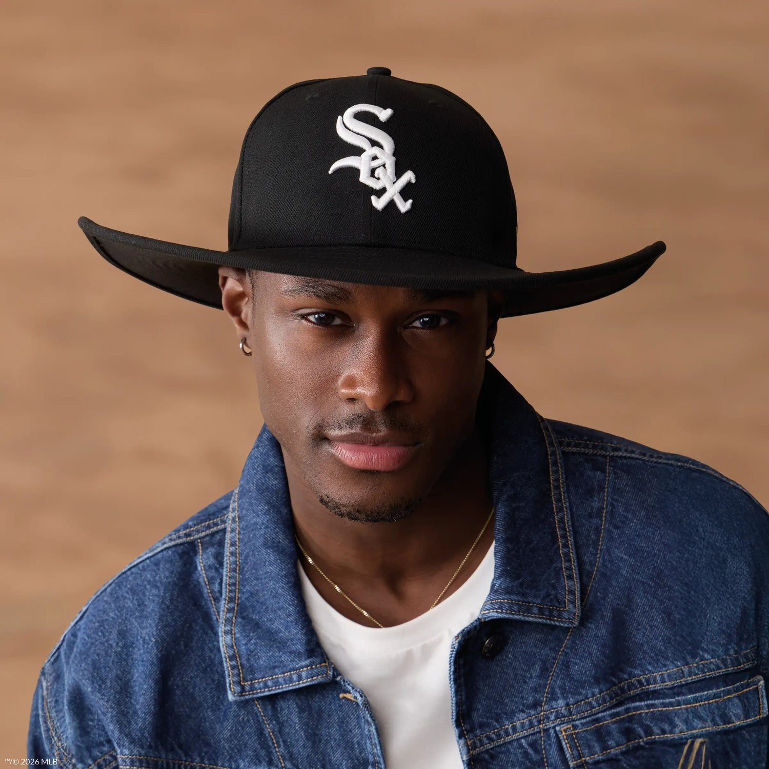 A man wearing a black cowboy-style baseball cap with a white embroidered 'Sox' logo, a blue denim jacket, and a white t-shirt, set against a warm brown background.