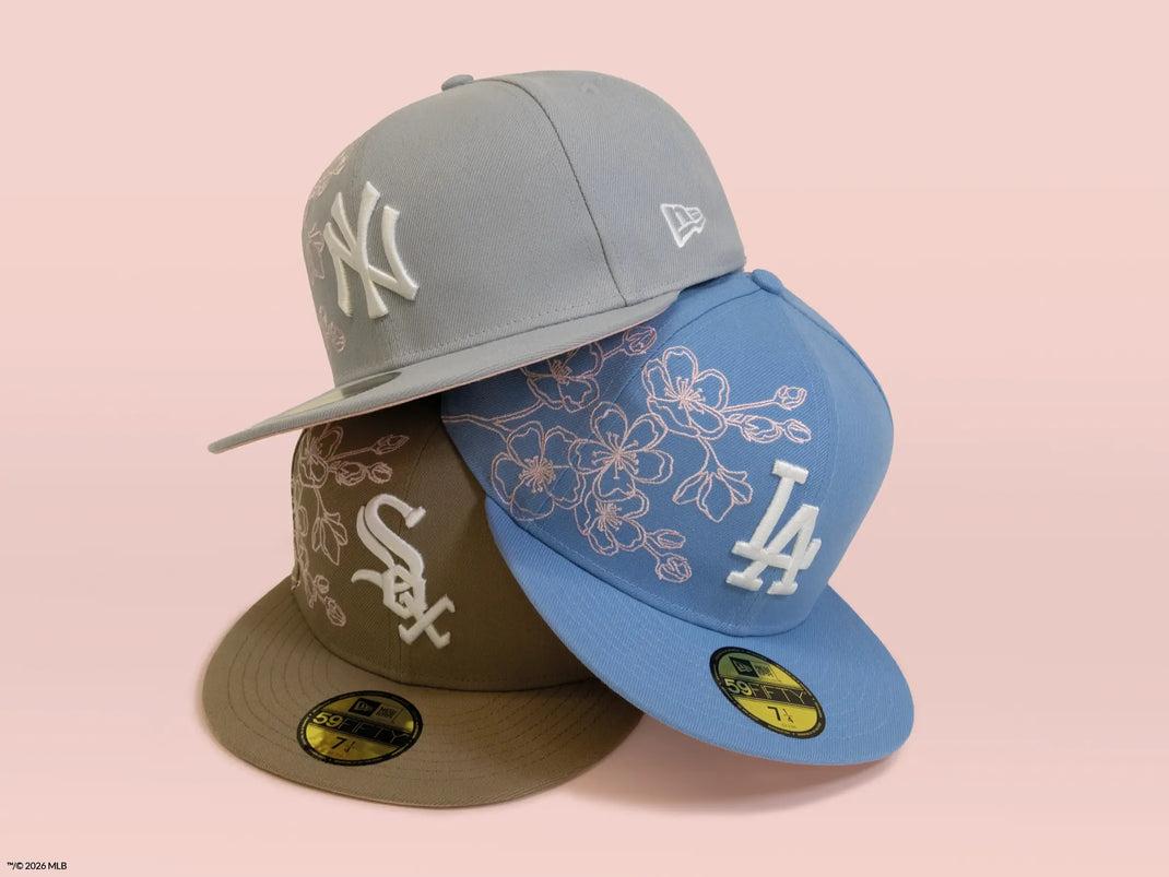 Three New Era 59FIFTY fitted baseball caps arranged in a fan shape on a soft pink background: a gray New York Yankees cap on top, a blue Los Angeles Dodgers cap in front, and a brown Chicago White Sox cap behind.