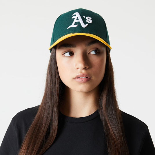 A girl wearing a green Oakland Athletics cap with a yellow brim and white embroidered 'A's' logo, paired with a black crewneck shirt against a light gray background.