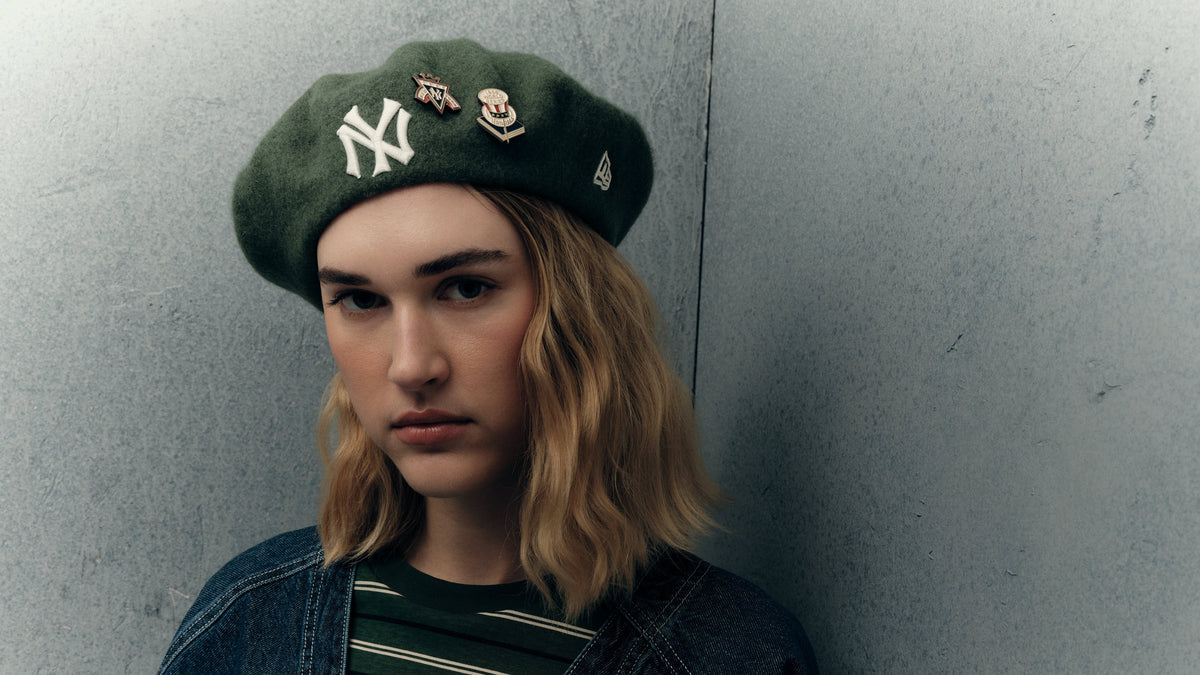 A girl, wearing a dark green New Era New York Yankees beret, a blue denim jacket, and a green and black striped t-shirt.