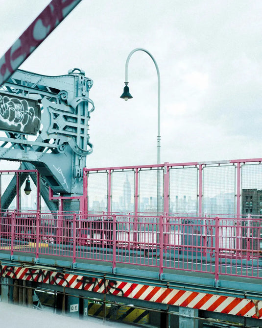 Part of a New York bridge with a pink railing and a streetlight against a gray sky.