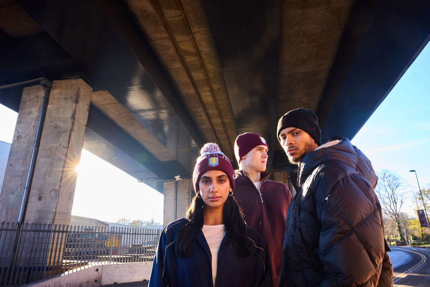 Three people standing under a bridge with a road and trees in the background wearing aston villa new era beanies