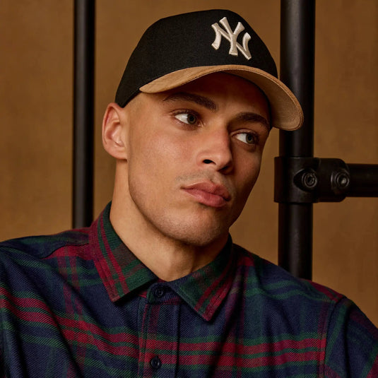 Man wearing a black cap with a logo and a plaid shirt against a brown background