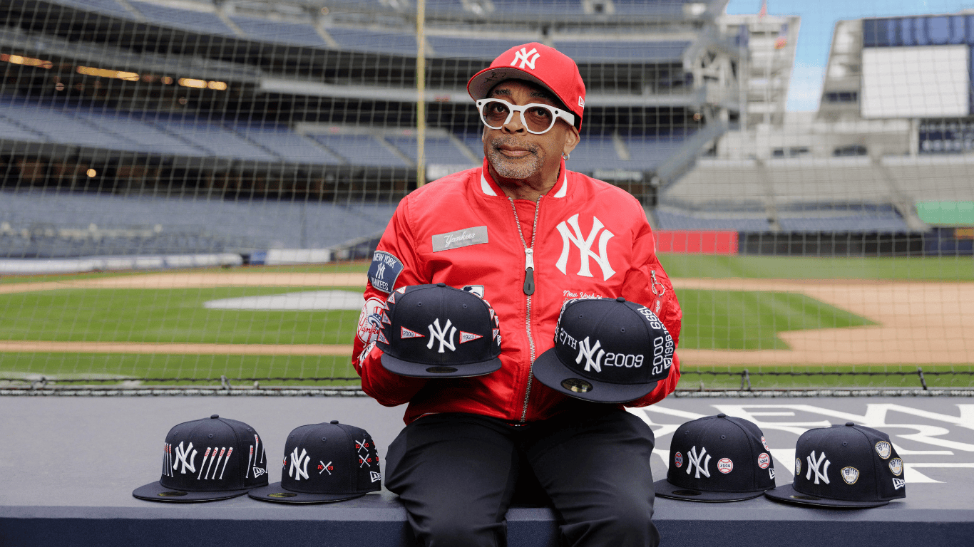 Spike Lee sitting at Yankees stadium holding New Era Caps
