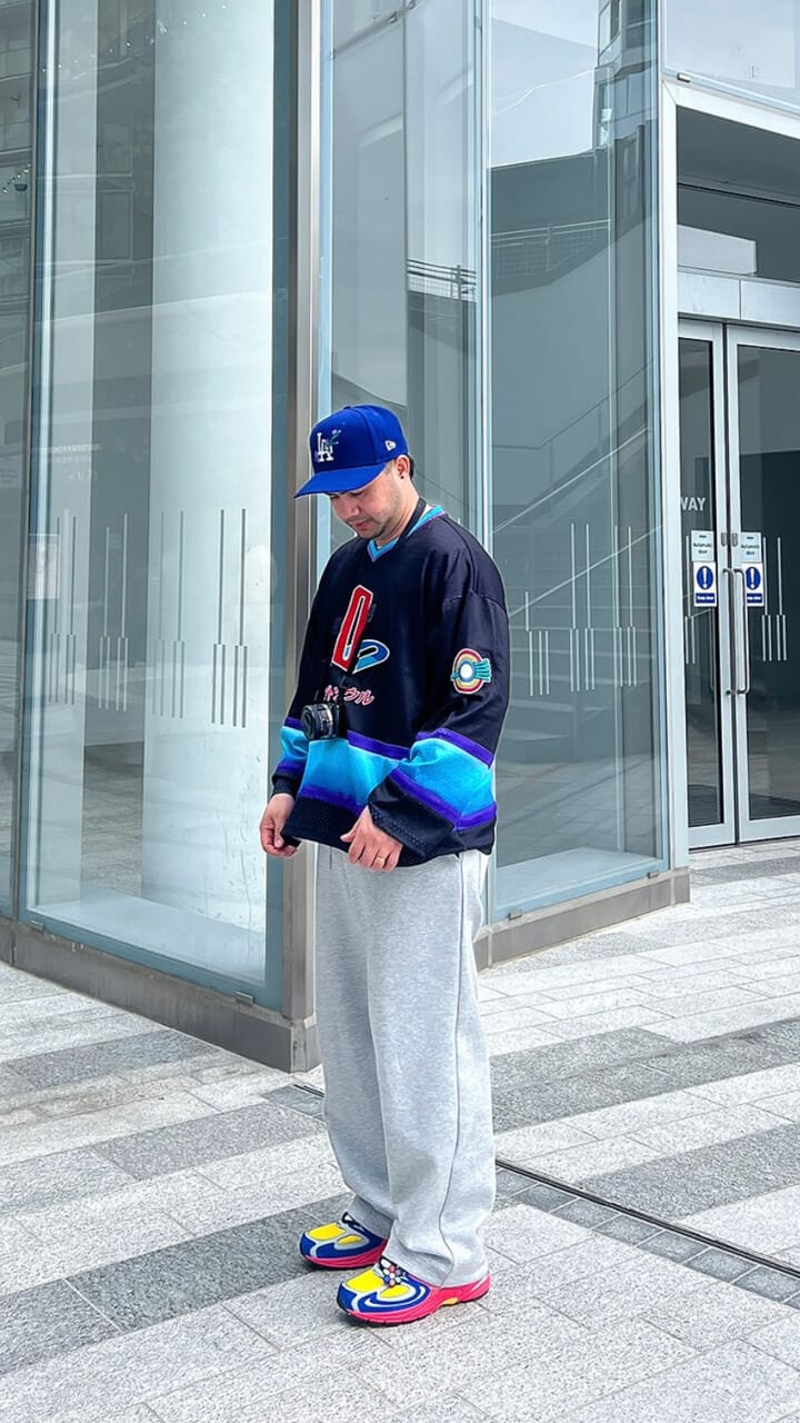 A man looking down at the floor wearing a blue LA Dodgers 59FIFTY cap, navy sweatshirt, grey joggers and yellow shoes.