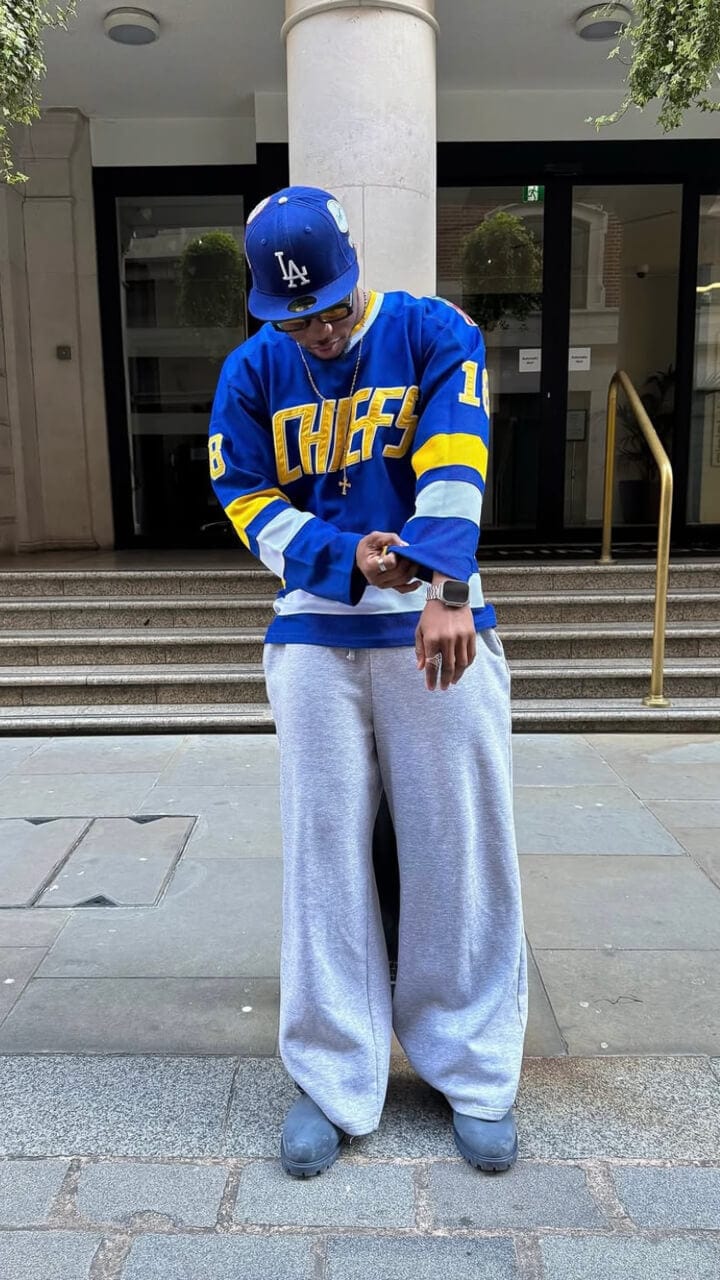 A man wearing a blue and yellow sweatshirt with 'Chefs' printed on it, grey pants, and a blue 59FIFTY LA Dodgers cap standing on a street.