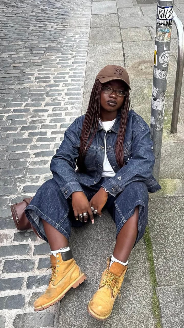 A women sitting down on the pavement wearing a brown New Era New York Yankees cap, matching denim outfit and nubuck Timberland boots. 
