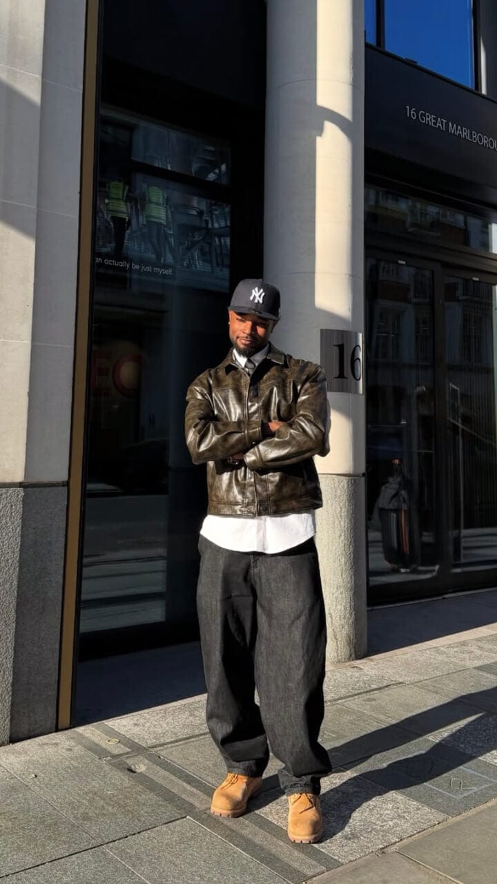 A man standing in front of a building with his arms folded  wearing a black 59FIFTY cap with a white New York Yankee logo, a green leather jacket, white t-shirt, denim jeans and nubuck timberland boots.