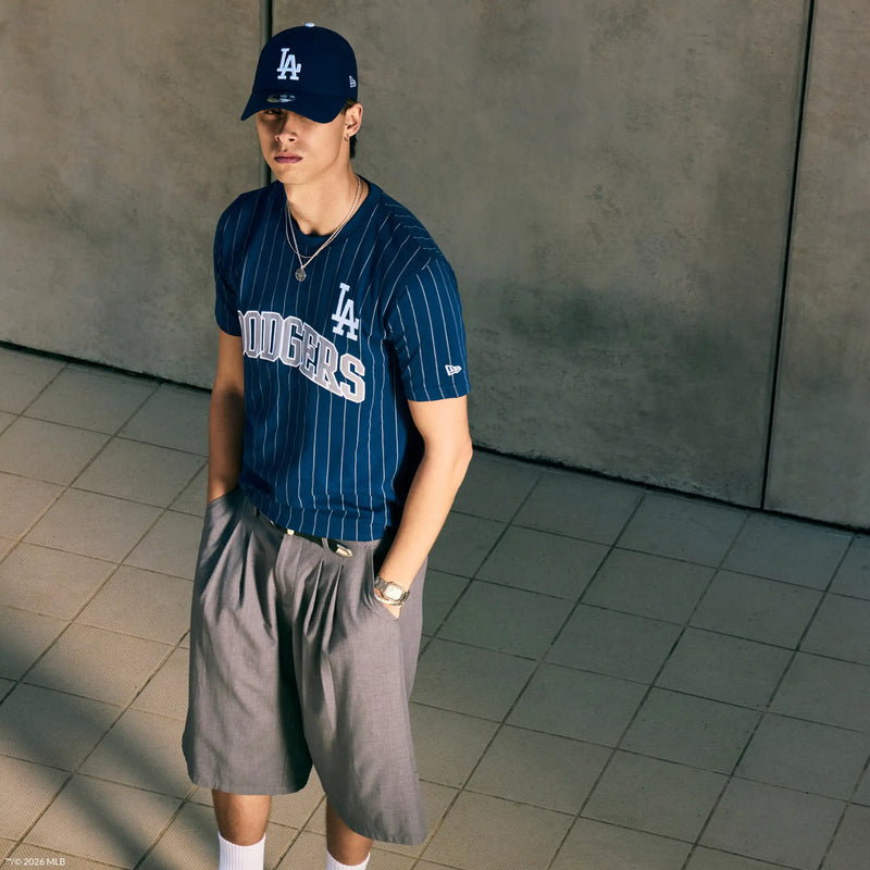 A man wearing a navy blue pinstriped Los Angeles Dodgers t-shirt, gray shorts, blue LA cap, and white socks, standing in a tiled indoor space with a concrete wall background.