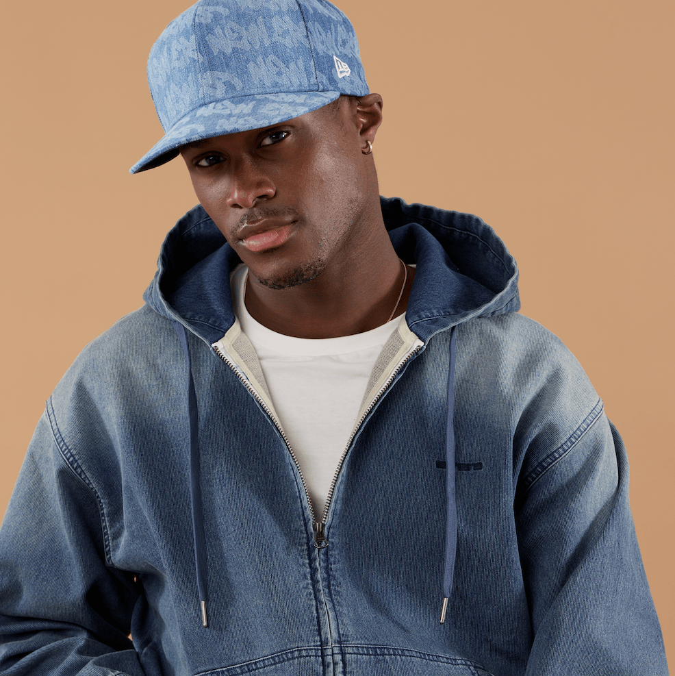 A man wearing a blue denim zip-up hoodie, a blue patterned New Era cap, and a white t-shirt, posed against a solid warm beige background.