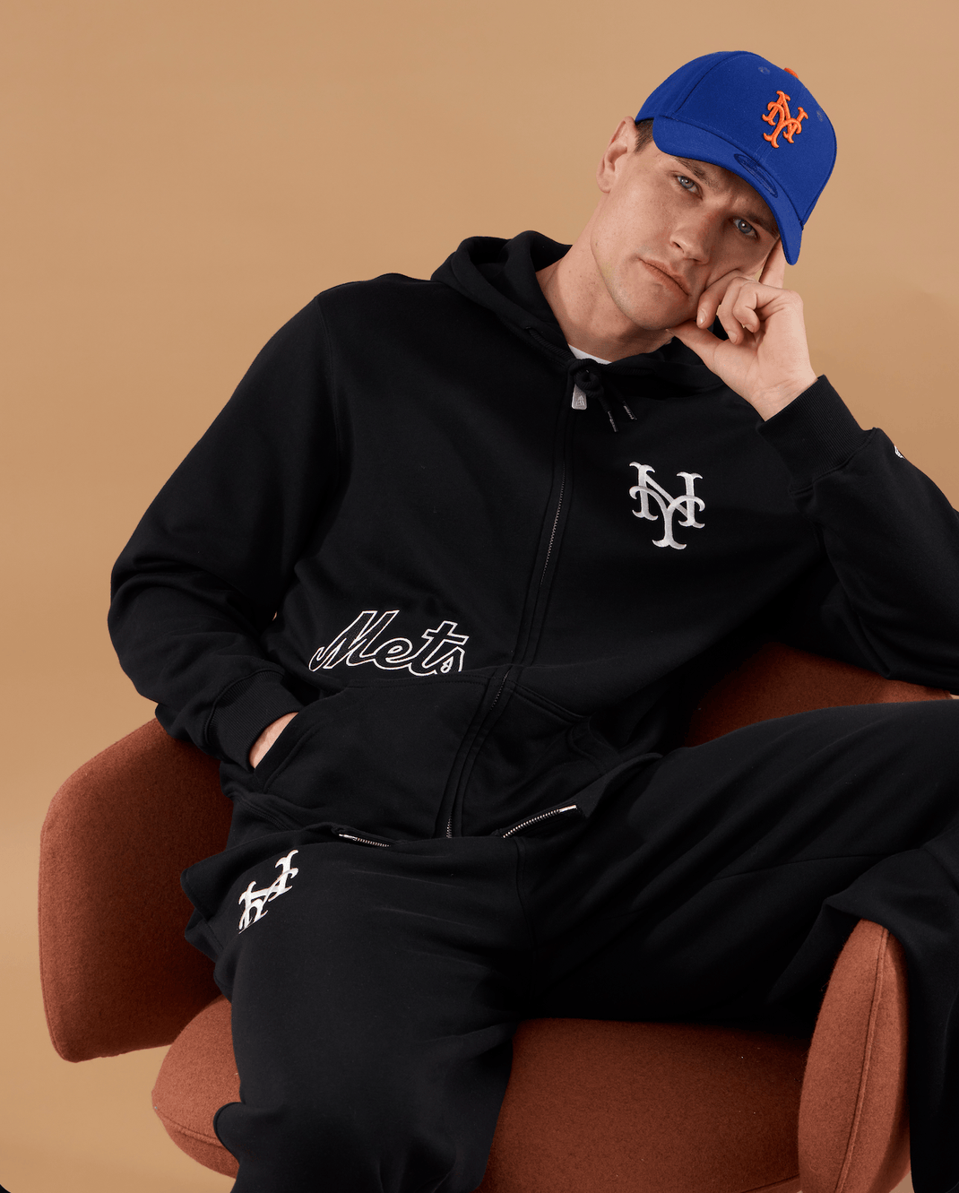 A man wearing a black zip-up hoodie and matching black sweatpants with white embroidered New York Mets logos, seated in a brown armchair against a warm beige studio background. He is also wearing a blue New York Mets baseball cap.