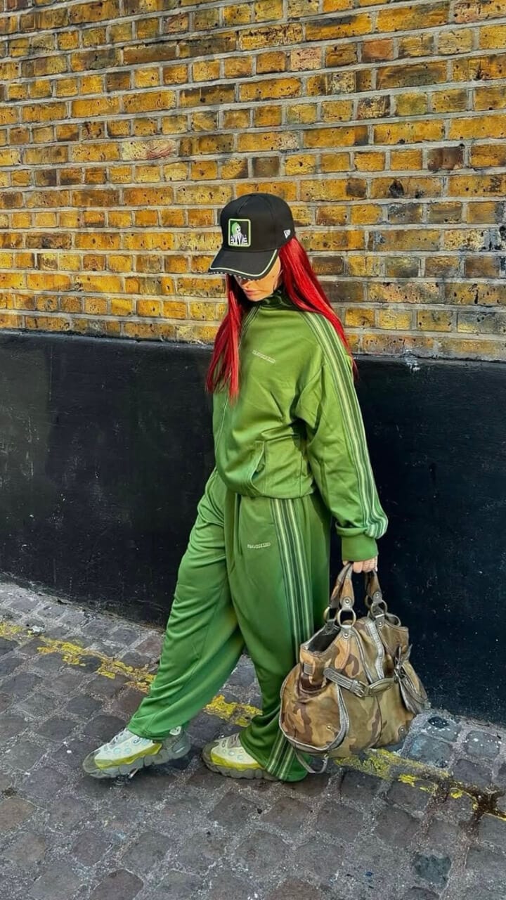 A women with red hair wearing a green tracksuit and black New Era Beetlejuice cap against a black and brick wall holding a camo duffle bag.