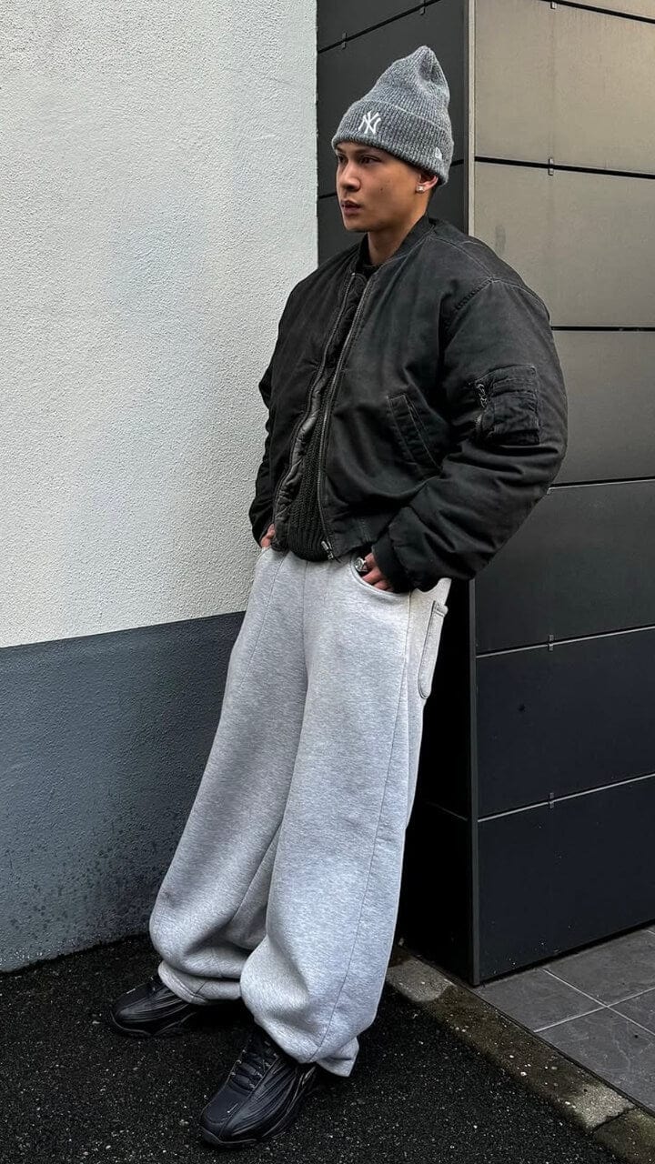 A man leaning on a black wall wearing a grey New Era New York Yankees beanie hat, black bomber jacket, grey joggers and black shoes. 