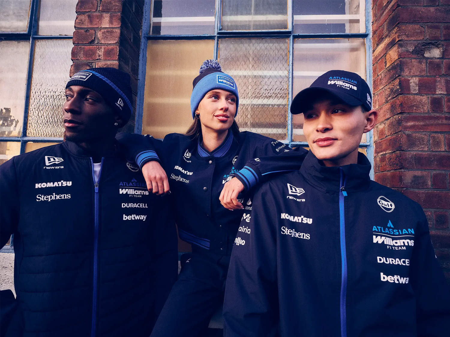 Three individuals wearing matching dark blue new era williams motorsports jackets and new era wiliams hats with various logos in front of a brick wall.